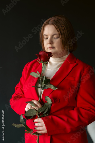 Stylish woman with closed eyes holds a red rose. She wears a vibrant red blazer against a dark background. This studio portrait evokes feelings of romance, serenity, and quiet contemplation