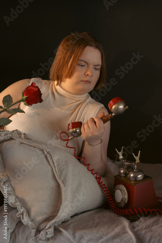 A retro styled portrait of a sad young woman with short hair lying in bed. She holds a red rose a vintage telephone, looking down with a pensive expression. Warm, moody lighting on a dark background