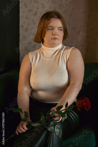 A thoughtful young woman with short red hair sits on a dark sofa holding a single red rose. She has a pensive, melancholic expression. Muted, moody lighting creates a cinematic and emotional scene