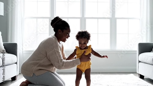 Happy African American mother with joyful mood watching her baby taking first steps against bright living room background