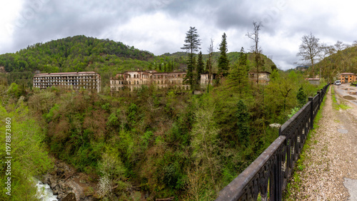 Panoramic view of abandoned residential blocks in mountain . Ornate bridge railing in the foreground overlooking a river valley and decaying soviet architecture in a ghost town. Akarmara, Abkhazia.