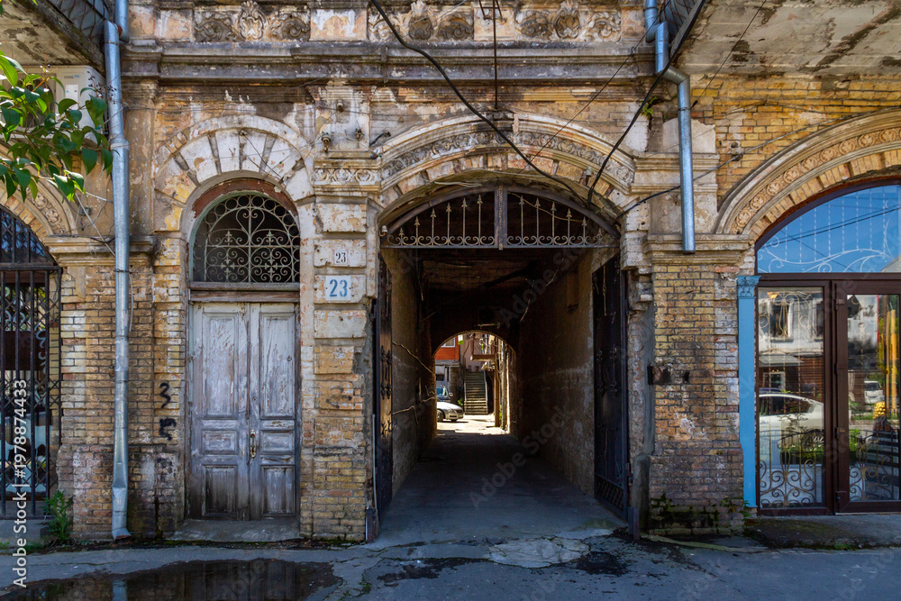 Fototapeta premium Classic architecture of an old brick building facade in Sukhum with an arched driveway and ornate decorations. Weathered wooden doors and vintage iron gates on a sunny spring day. Abkhazia.