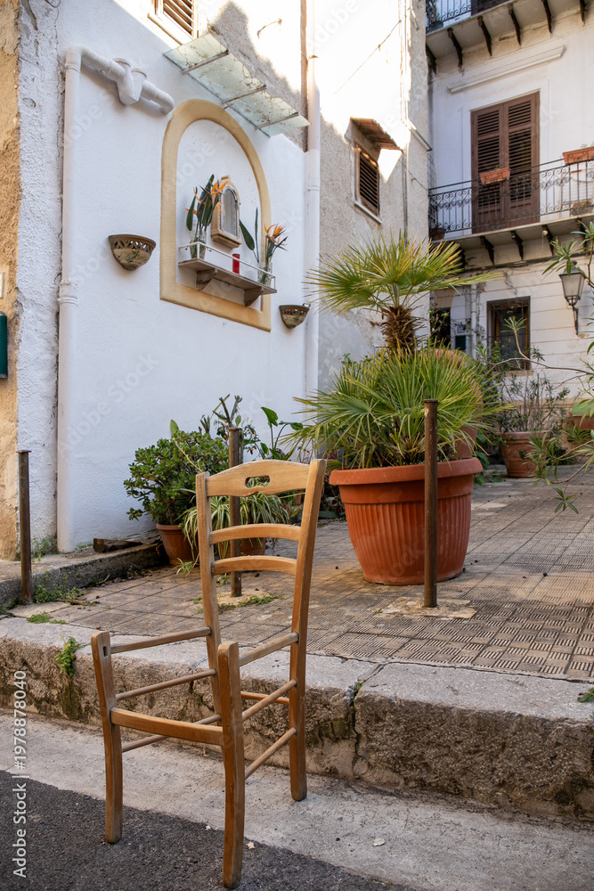 Obraz premium Charming quiet alleyway in a historic village in Sicily featuring whitewashed stucco walls, a small decorative shrine niche, terracotta potted plants, and a simple wooden chair