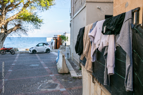 Coastal street scene, likely in Italy, featuring laundry air-drying on a fence overlooking the sea, with a white hatchback car and a motorcycle parked nearby, capturing traditional Mediterranean life