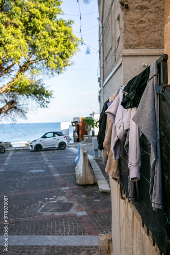 Coastal street scene, likely in Italy, featuring laundry air-drying on a fence overlooking the sea, with a white hatchback car and a motorcycle parked nearby, capturing traditional Mediterranean life
