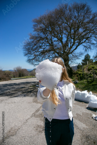 Person holding a large lump of compacted snow or ice in front of their face on a sunny outdoor road, with a tree in the background, capturing a playful winter moment, natural texture