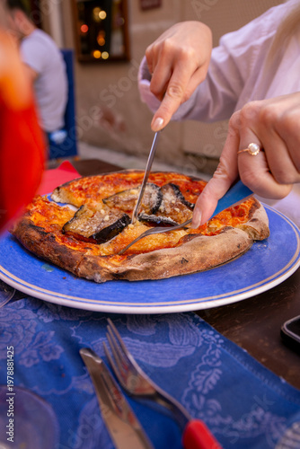 Person cutting a slice of Neapolitan-style pizza topped with eggplant, served on a blue plate, capturing a casual Italian restaurant dining experience with traditional cuisine, fresh ingredients