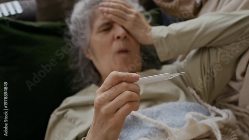 Medium selective focus shot of elderly Caucasian female seasonal flu sufferer lying on sofa covered with blanket, staring at thermometer with hand on forehead, coughing and sneezing