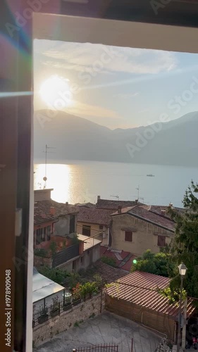 Beautiful sunset view from a window overlooking Lake Garda, the Alps and a small Italian-style village. Warm light over a scenic mountain and lakeside landscape.