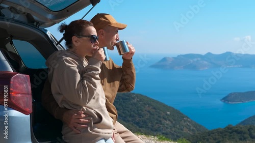 Romantic couple drinking coffee on a road trip admiring the seascape. Romantic couple sitting in the trunk of their car, drinking coffee from steel mugs and admiring the beautiful seascape
