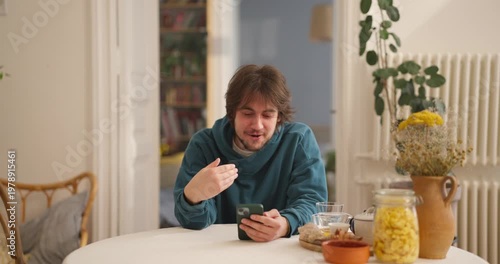 A young man enjoys moments at a table in his cozy kitchen, sipping and smiling with his phone in hand, sharing thoughts