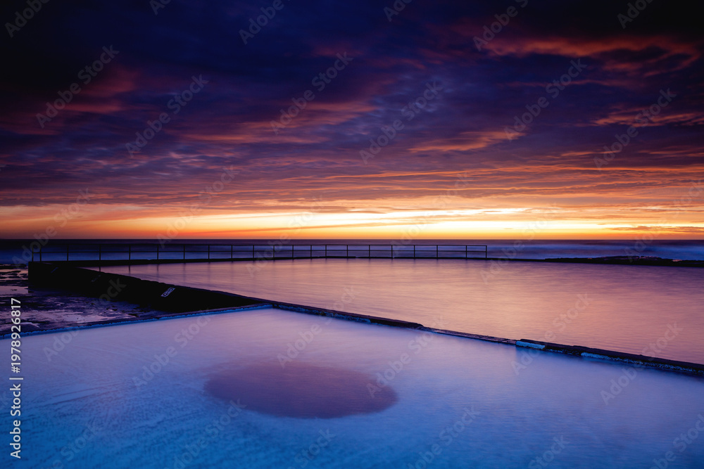 Obraz premium A beautifully stunning, vibrant crimson sunrise above the Wombarra Rock Pools located on the beach in the seaside village of Wombarra, on the Illawarra coastline. Captured in the golden hour.