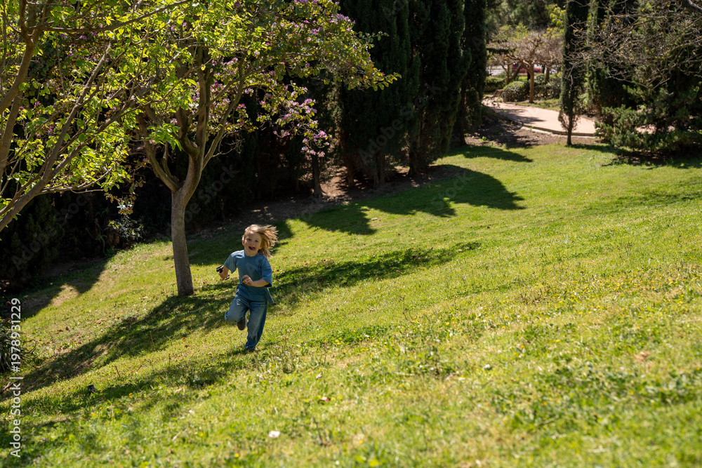 Obraz premium Little blond boy running on green hill in sunny park, childhood freedom concept, outdoor activity, joyful movement, spring nature, lifestyle scene, carefree play, landscape background