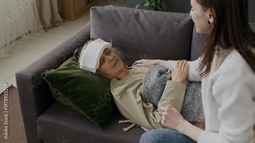 Medium shot of young Caucasian granddaughter putting cold towel compress on forehead for grandmother sick with flu, lying on couch with eyes closed, touching hand with gratitude and love
