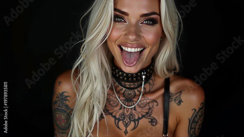 A woman with long blonde hair is laughing and sticking out her tongue. She is wearing a black choker with silver studs and a black spaghetti strap top.