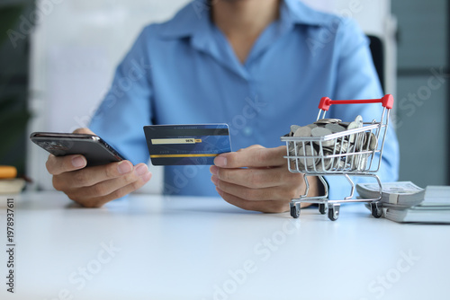 Person using smartphone and credit card for online shopping with stack of cash and coins in shopping cart.
