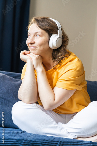 Tableau sur toile Woman in a yellow shirt and white pants sitting comfortably on a sofa, smiling while listening to music with headphones