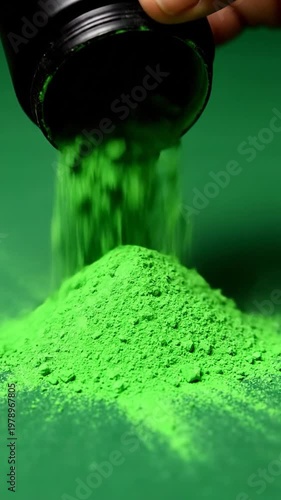 A hand pours vibrant green cosmetic powder from a small black container onto a matching surface