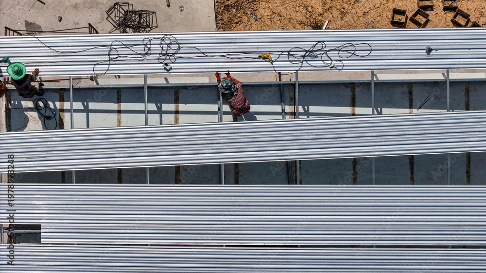 Naklejka premium Aerial view of construction workers installing roofing panels on a building, surrounded by tools and materials, showcasing teamwork, precision, and the construction process in progress.