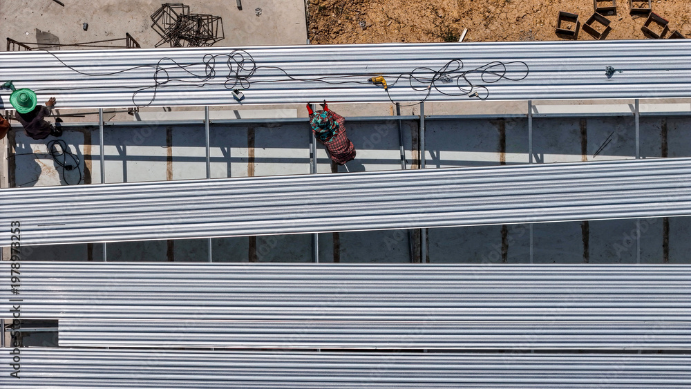 Naklejka premium Aerial view of construction workers installing roofing panels on a building, surrounded by tools and materials, showcasing teamwork, precision, and the construction process in progress.