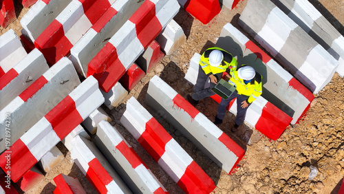 Obraz na płótnie Close up top view of two construction workers in safety gear using a laptop at a worksite, surrounded by red and white barricades, emphasizing teamwork, planning, and modern construction technology