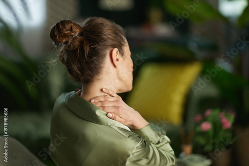 Fotografie A woman in a green blouse reaches back to touch her neck, depicting muscle tension and the need for a mindful pause and physical relief in her home environment