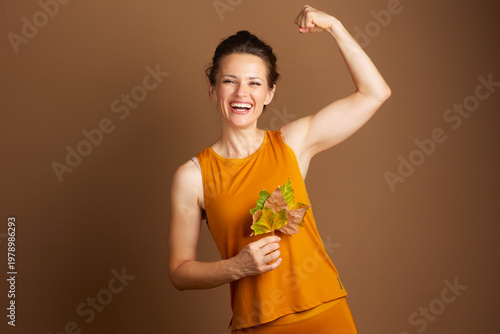 Canvastavla An adult woman in mustard yellow activewear laughs and flexes her bicep while holding autumn leaves, embodying strength, vitality, and cheerful seasonal health
