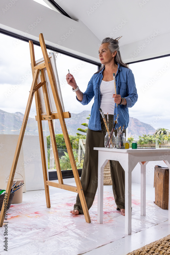 Naklejka premium Senior female artist standing barefoot in bright studio, painting on wooden easel above drop cloth