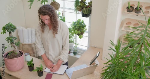 Vertical video: Opening notebook owner in beige top noting at counter checking plant using laptop