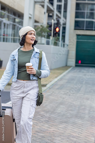 Asian adult woman walking on city plaza pulling rolling suitcase holding paper cup