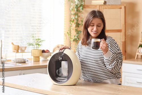 Young woman with cup and coffee machine at table in kitchen