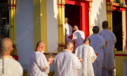 Принти на полотні Thai men in white walk in line with palms together carrying folded orange monk robes as medium to large group approaches temple doorway, warm light before ordination