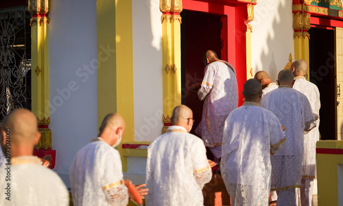 фотографія Thai men in white walk in line with palms together carrying folded orange monk robes as medium to large group approaches temple doorway, warm light before ordination