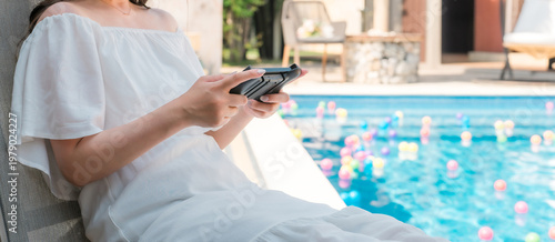 Принти на полотні A young Asian woman sits in a recliner chair by the poolside playing a game
