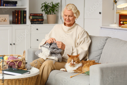 Senior woman with ginger cat knitting sweater on sofa at home