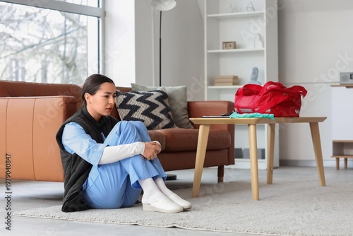 Exhausted female healthcare worker in scrubs sitting on living room floor aft...