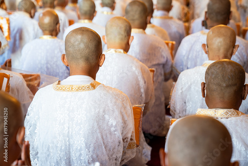 Obraz na płótnie A rear perspective shows white clad ordinands seated in rows inside the temple holding orange robes with shaved heads and folded hands in a soft blurred ritual setting