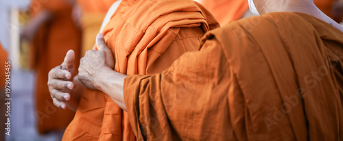 A senior monk adjusts a novice robe as hands tighten the woven belt around orange cloth in sunlight with focus on texture folds and precise fitting.