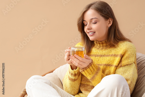 Young woman with glass cup of green tea in armchair on beige background