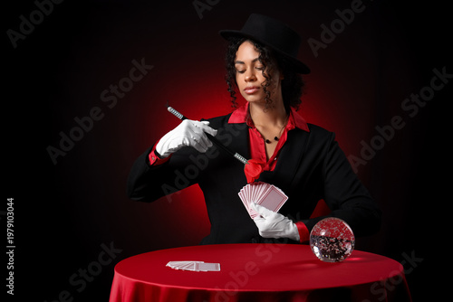 Картината върху платно Young African-American magician with magic wand, cards and crystal ball on table