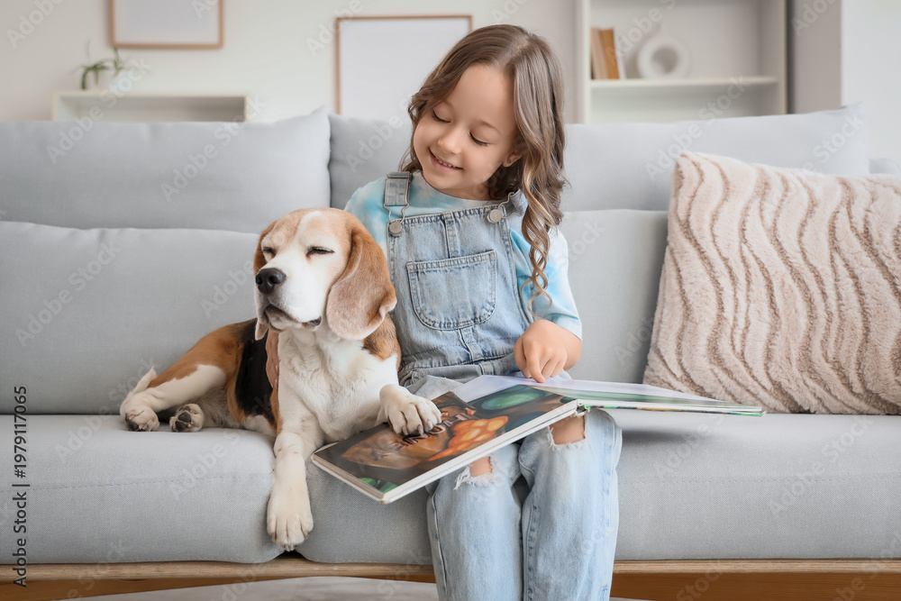 Fototapeta premium Cute little girl with Beagle dog reading book on sofa in living room
