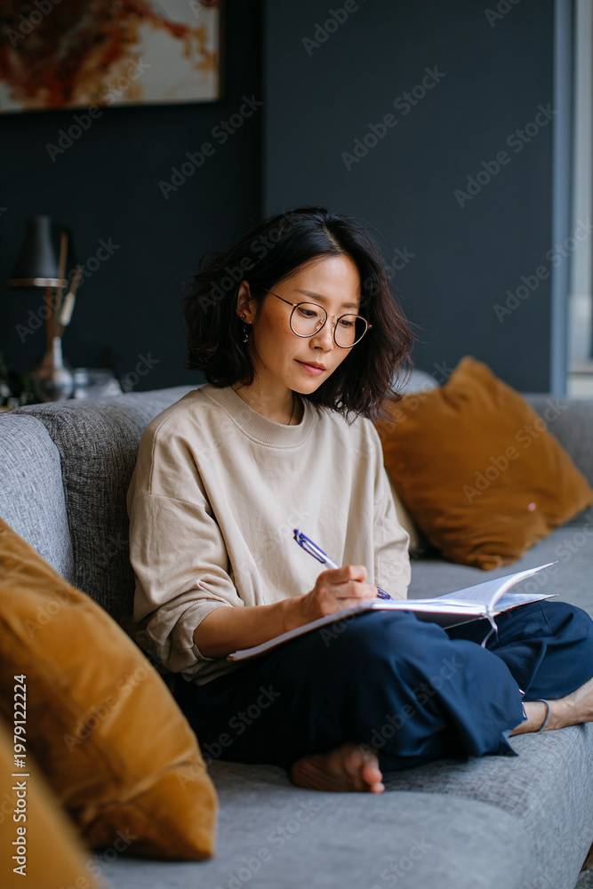 Obraz premium An Asian woman is sitting on a sofa, working with a notebook beside her.