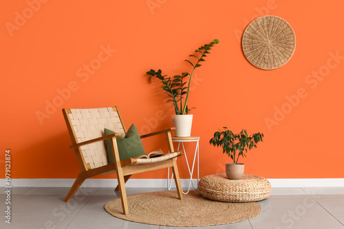 Comfortable armchair with wicker pouf and houseplants near orange wall