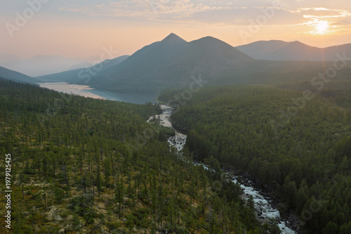 Summer evening mountain landscape. Aerial view of the river, lake, mountains and taiga. The sun over the mountains. The source of the Kuel-Sien River from Lake Dancing Grayling, Magadan Region, Russia