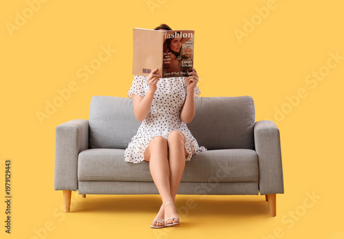 Fényképezés Beautiful young woman reading magazine on sofa against yellow background