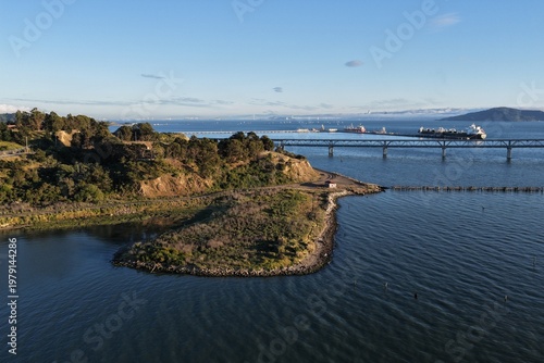A drone view captures Point Molate Beach Park near golden hour, where warm light settles over the shoreline, gentle waves meet the sand, and the bay reflects soft evening tones