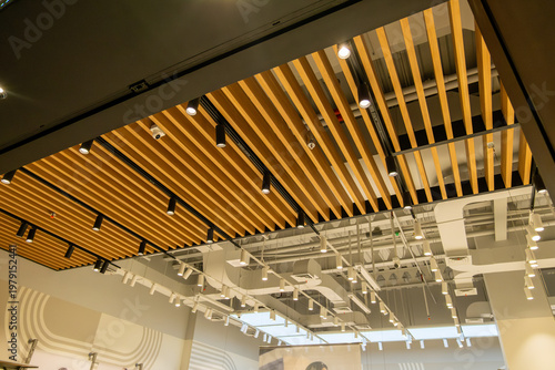 Modern commercial interior ceiling featuring wooden slats, exposed industrial ducts, hanging LED lights, CCTV security camera, and ventilation systems in a contemporary retail space.