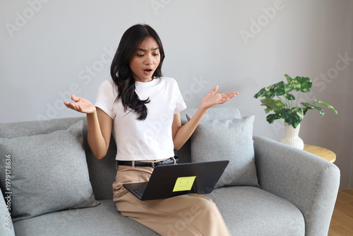 Fotografie A young woman sits on a sofa in her living room, anxiously staring at a laptop screen, reacting to the bad and unexpected news with frustration and anger, shrugging her shoulders and hands