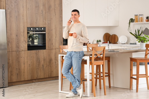 Handsome young man with cup of coffee talking by phone in kitchen