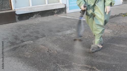 Man in airtight suit sprays disinfecting liquid chemicals antiseptic on street in city on day. Sanitation workers clean pavement, crosswalk. Sanitary measures in public place. Coronavirus pandemic